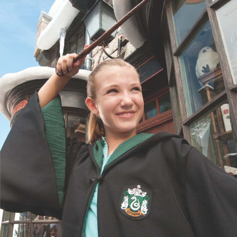 Una niña vestida con una toga de Hogwarts, apunta su varita mágica hacia el cielo. 