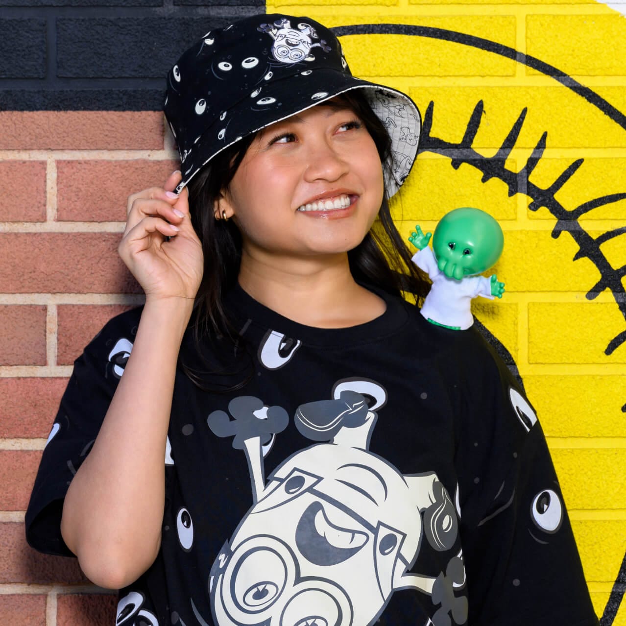 A smiling Guest in a Minions & Monsters-themed black shirt and hat with a small Goomi character figure on her shoulder.