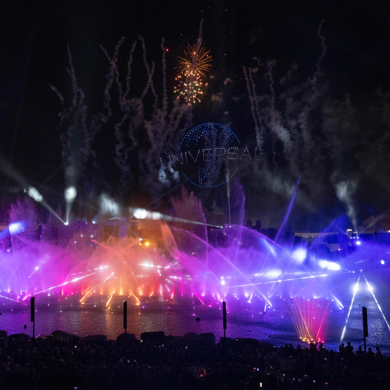 Un espectáculo nocturno con fuentes multicolores en el agua y fuegos artificiales, y el globo de “UNIVERSAL” iluminado en el cielo.