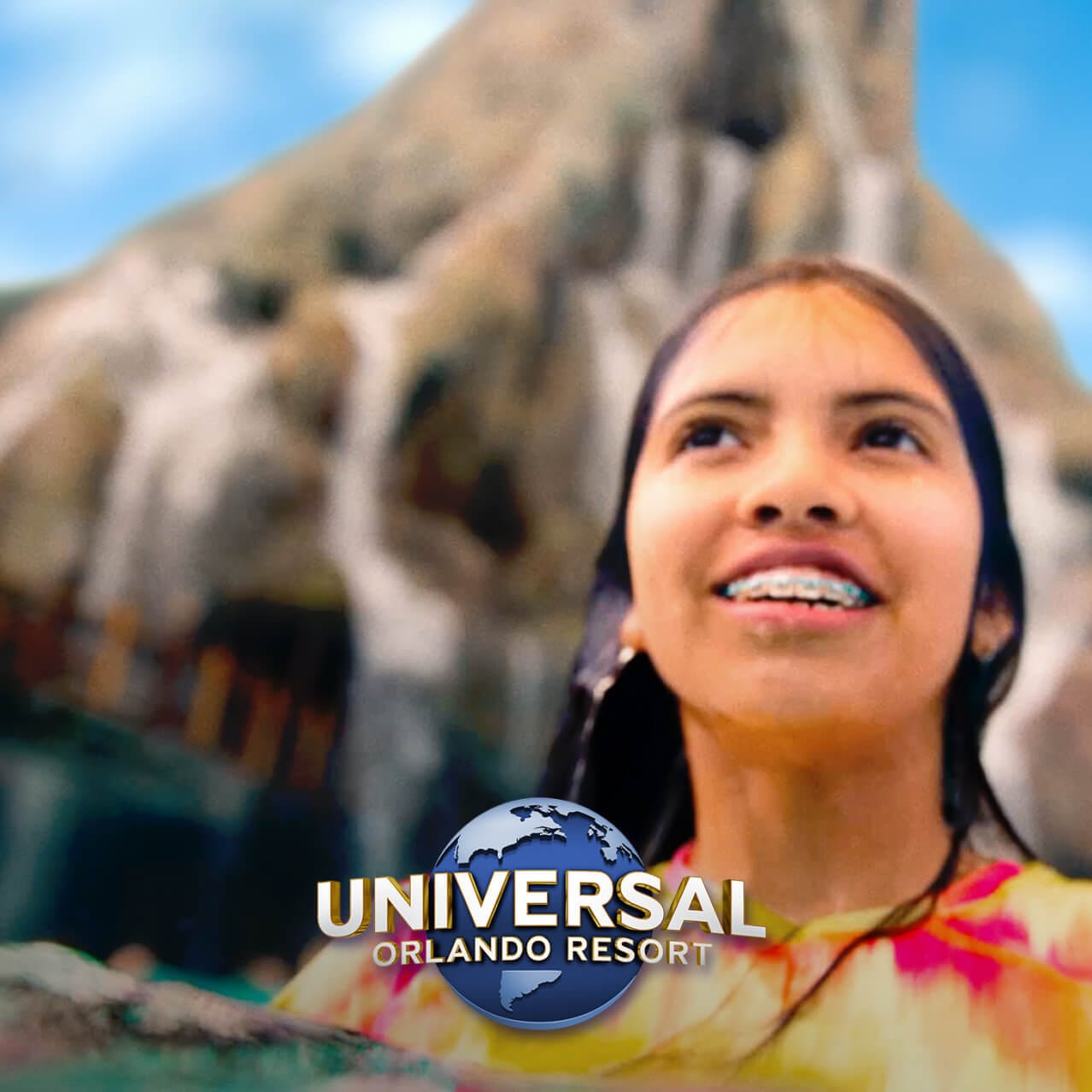 Una niña sonriente con el cabello mojado y aparatos ortopédicos mira hacia la distancia mientras el volcán del parque acuático se eleva en el fondo.