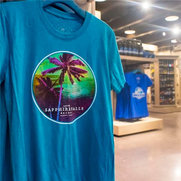 Una camiseta azul con una imagen de una palmera y un océano y las palabras "Loews Sapphire Falls Resort" exhibida en la tienda de regalos del hotel, Universal Studios Store™. 