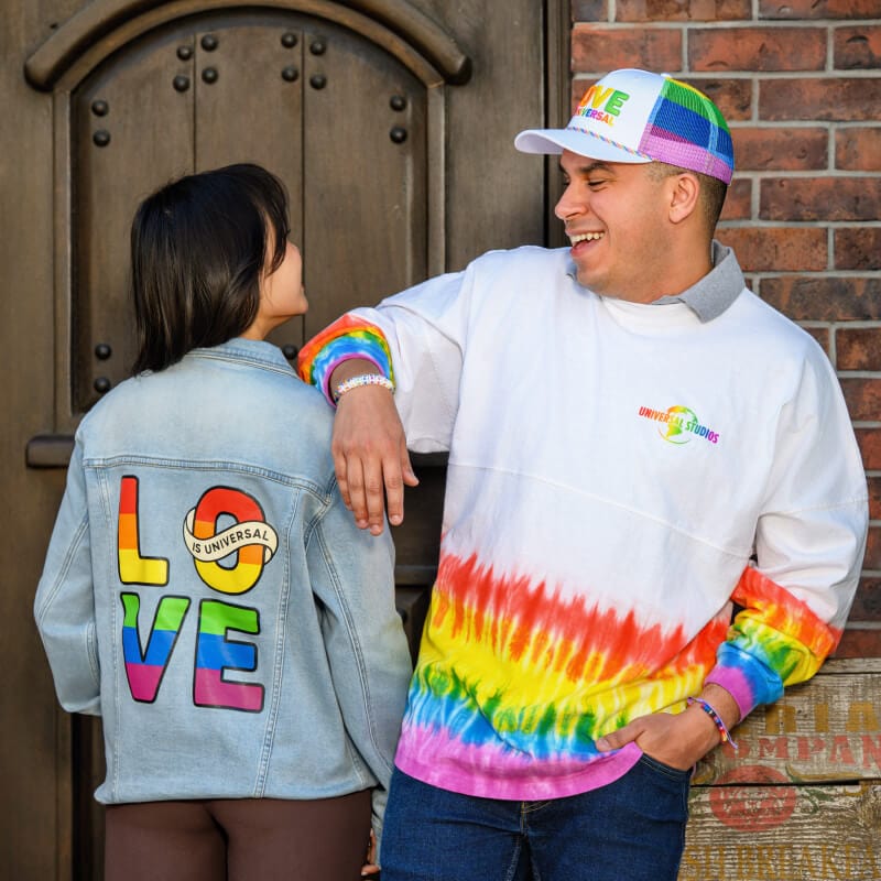 Una pareja con una chaqueta, una camiseta y un sombrero con el logotipo "LOVE IS UNIVERSAL" en los colores del arcoíris.
