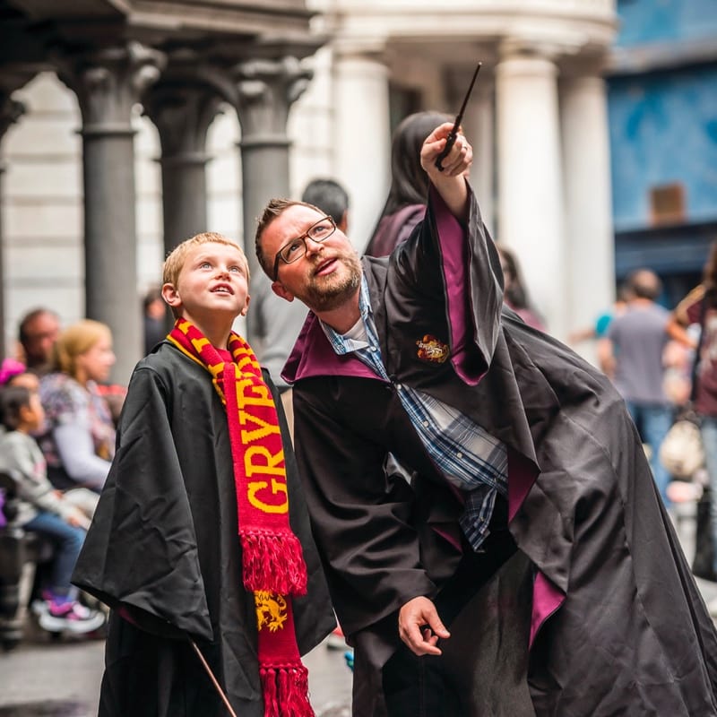 Un hombre y su hijo usan capas de Hogwarts mientras el padre apunta al aire con su varita.