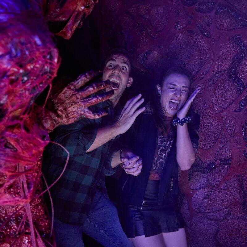 Una pareja que se toma de la mano y grita aterrorizada mientras una criatura viscosa les tiende su mano en descomposición en la oscuridad.