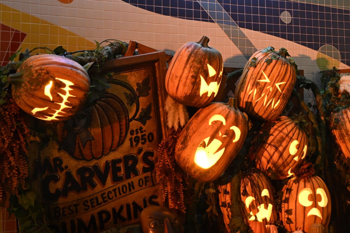 A variety of glowing carved jack-o’lanterns piled up next to a wooden “MR. CARVER’S BEST SELECTION OF PUMPKINS” sign.