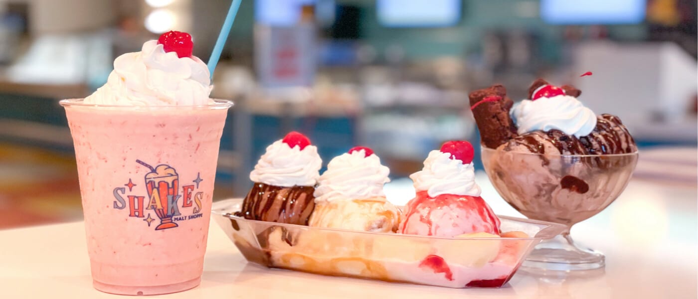 Shake de morango, banana split e sundae de chocolate em cima de uma mesa com um balcão de shakes ao fundo