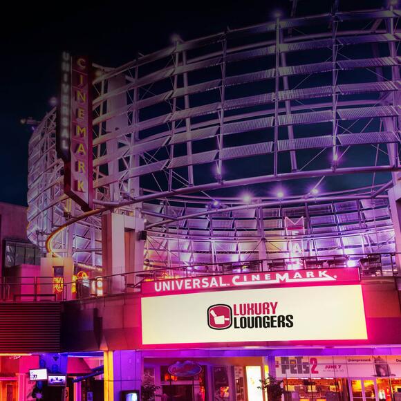 No Universal CityWalk, um letreiro brilhante de "BEM-VINDO AO UNIVERSAL ORLANDO RESORT" aparece ao lado da placa "POLTRONAS DE LUXO NO UNIVERSAL CINEMARK” que fica logo abaixo de um letreiro vertical do "UNIVERSAL CINEMARK" ao lado de "BK WHOPPER BAR" e placas do "PANDA EXPRESS" visíveis em segundo plano.