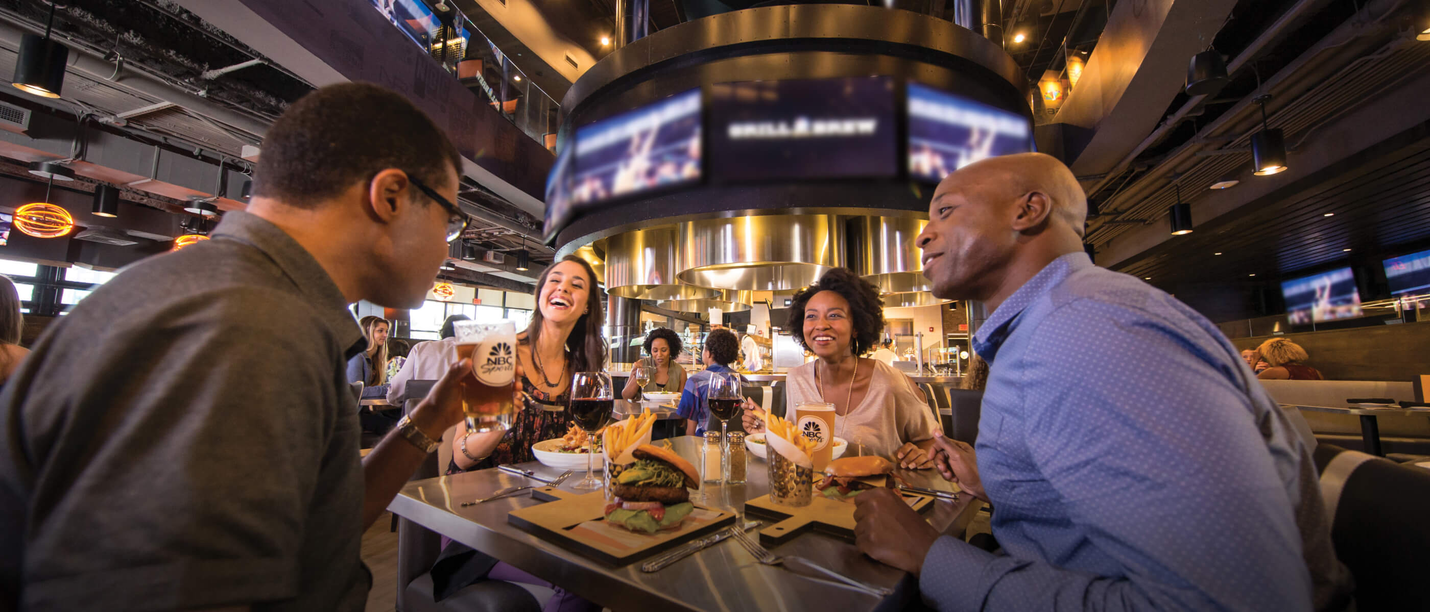 Four friends smiling and talking together over burgers, fries, beer, and wine at a table inside NBC Sports Grill & Brew, with large television screens visible in the background.