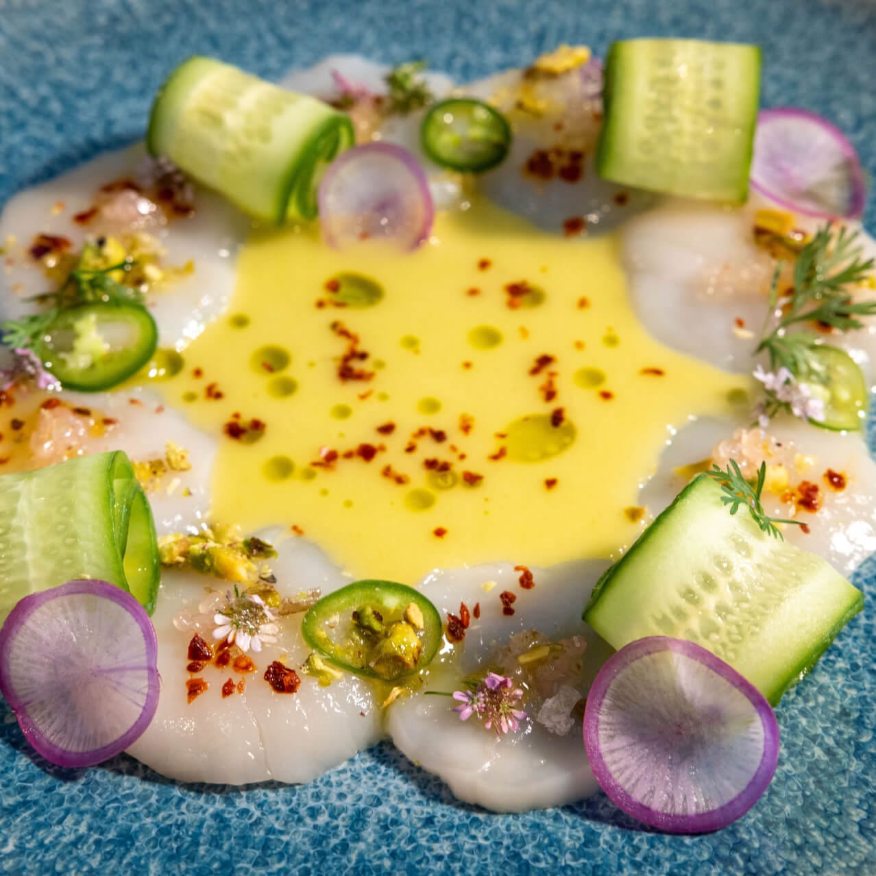 Scallop Crudo com vieiras frescas e cruas em um molho de laranja azeda e vinagre, harmonizado com fatias de pepino persa, pimenta Aleppo, pimentas serrano frescas e pistache crocante, servido no Bar Helios.