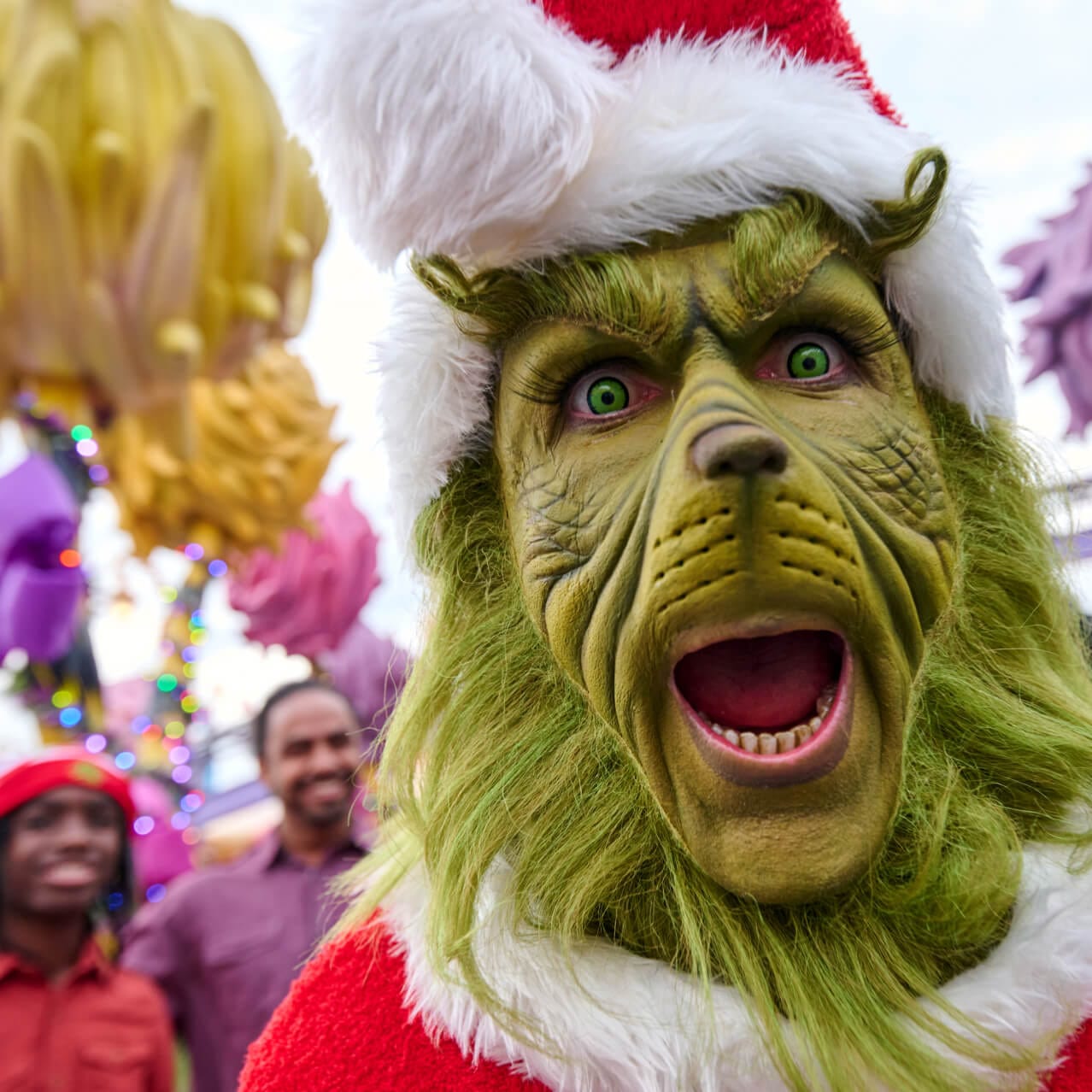 O Grinch vestido de Papai Noel com uma expressão animada e pessoas ao fundo.
