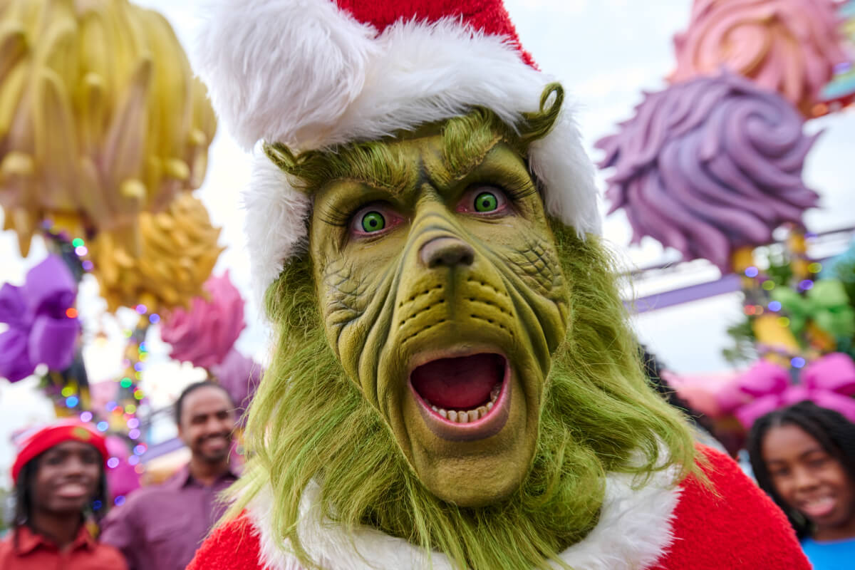The Grinch dressed as Santa making an excited expression as people stand in the background.