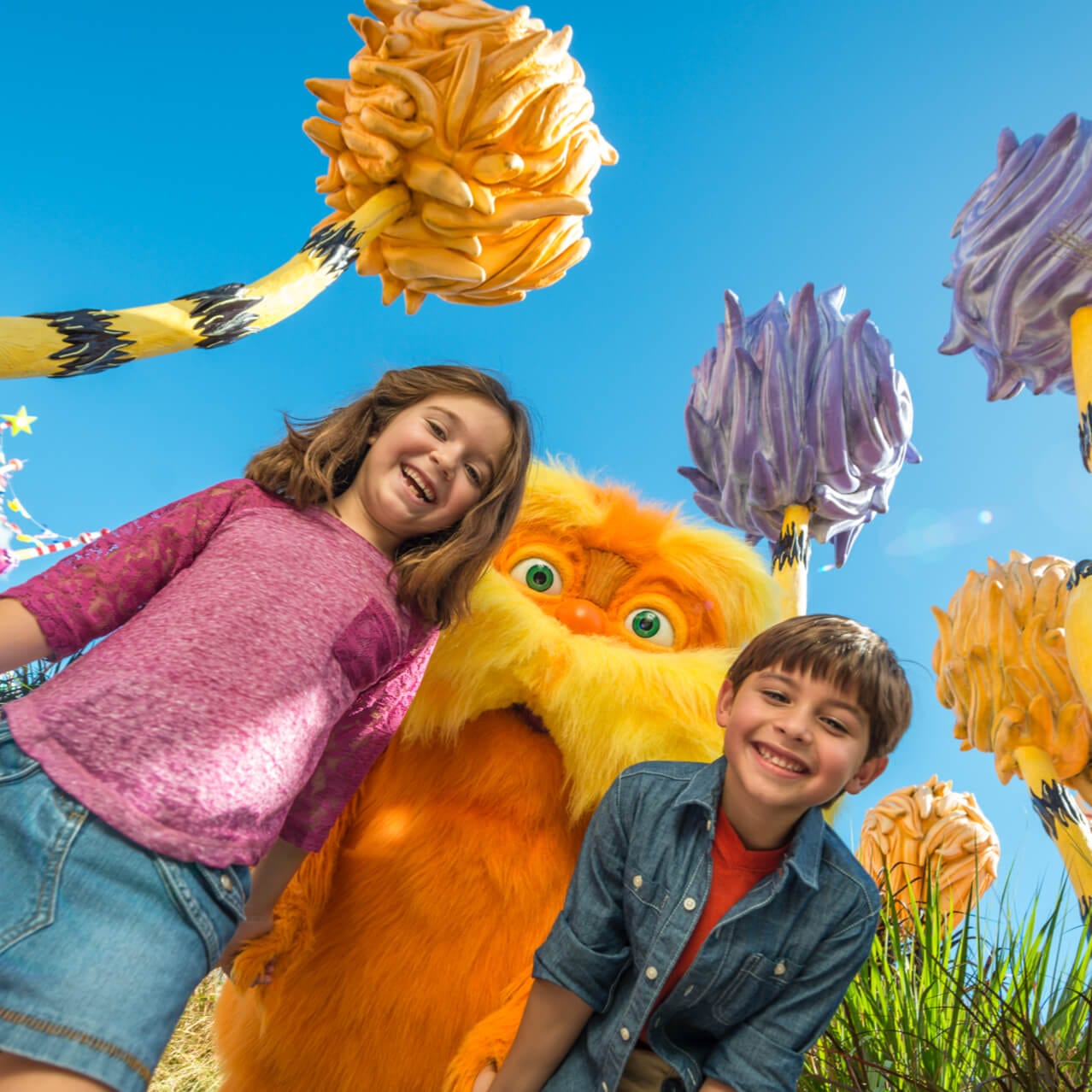 Duas crianças sorriem com o personagem laranja Lorax posando entre elas no Local com Personagens do Dr. Seus do Universal’s Islands of Adventure.