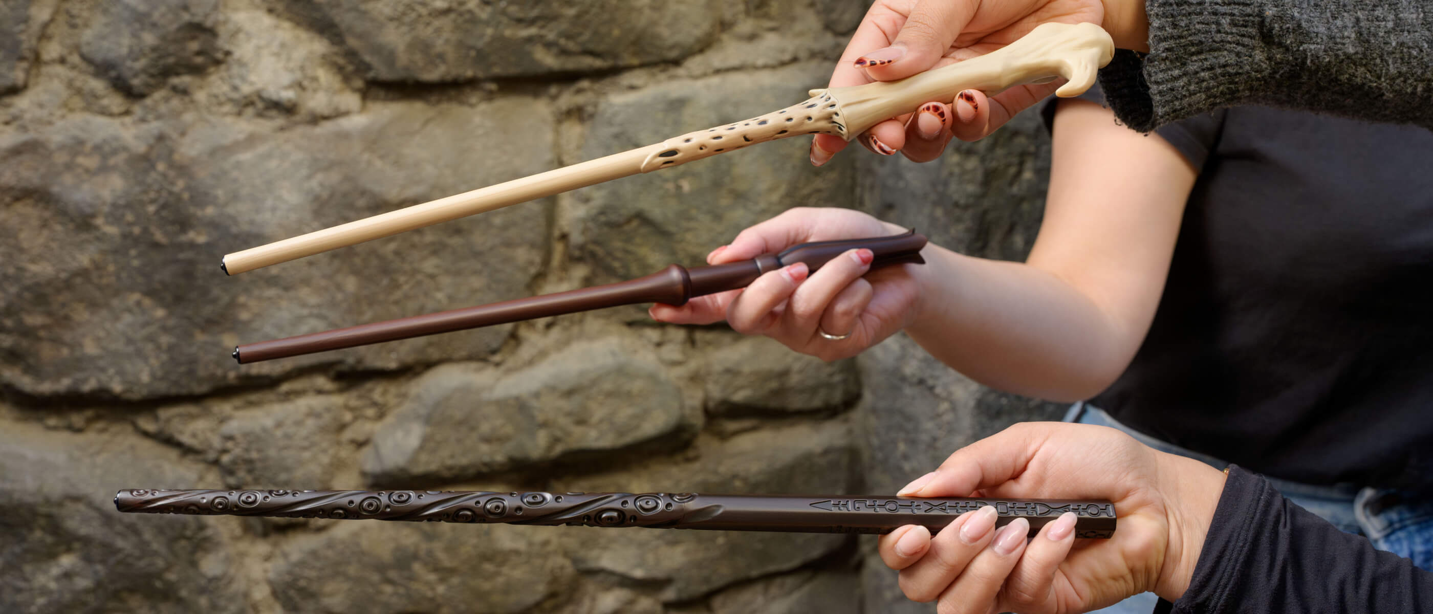 The hands of three Guests holding three different magic wands out in front of a stone wall, displaying their varying look