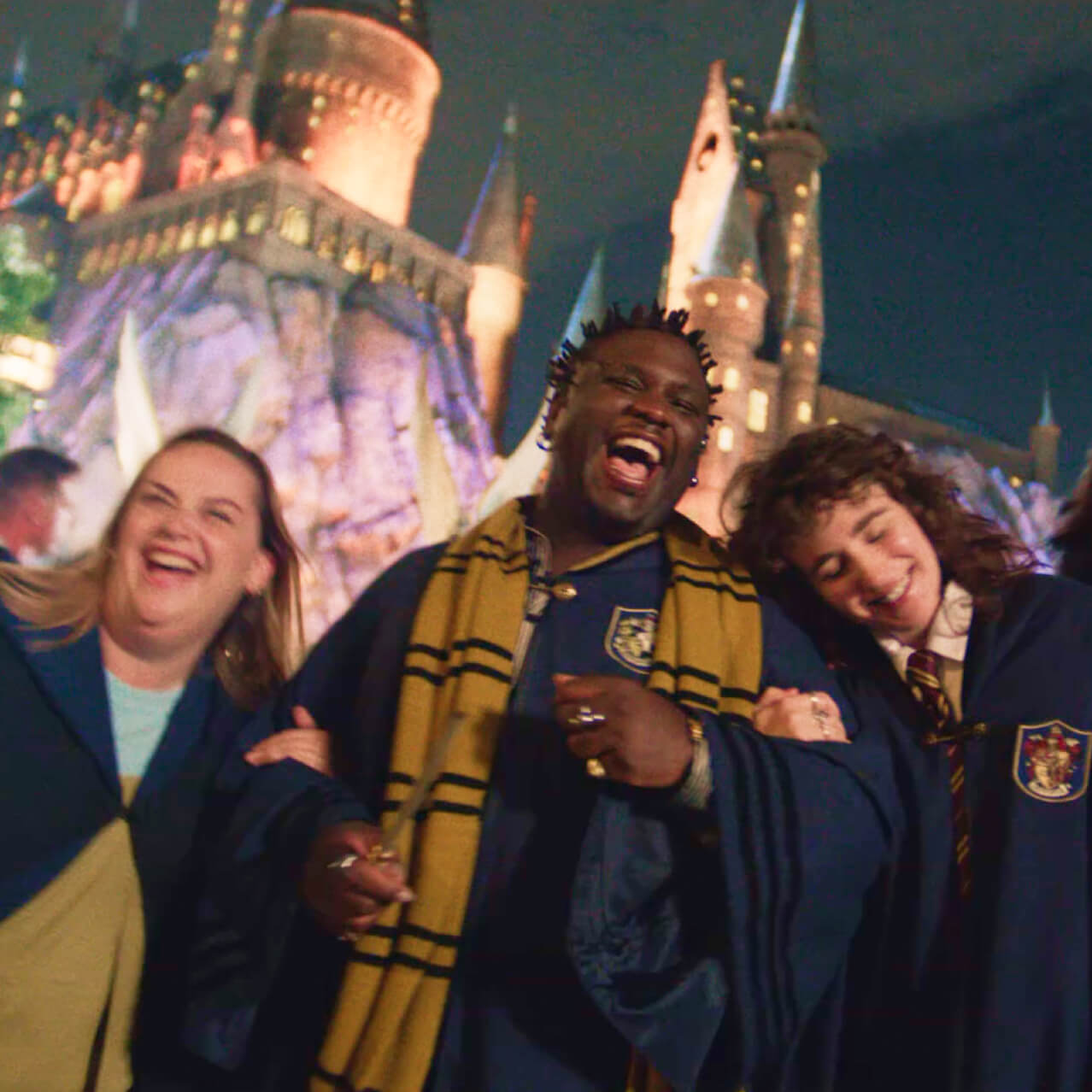 Amigos usando vestes de bruxo, sorrindo e rindo à noite, enquanto o castelo de Hogwarts iluminado se ergue ao fundo.
