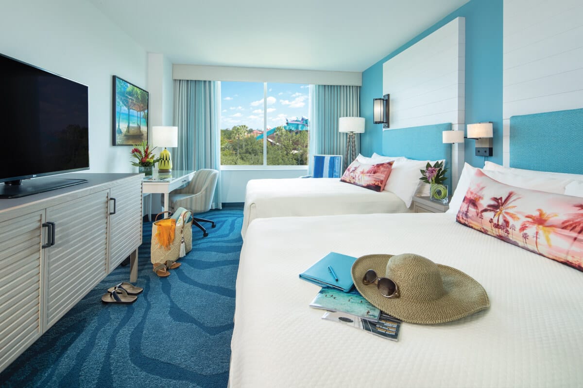 Um chapéu de sol e um óculos de sol estão sobre a cama em um elegante quarto branco e verde-água do Loews Sapphire Falls Resort no Universal Orlando, com duas camas queen size, uma escrivaninha e uma TV de tela plana.