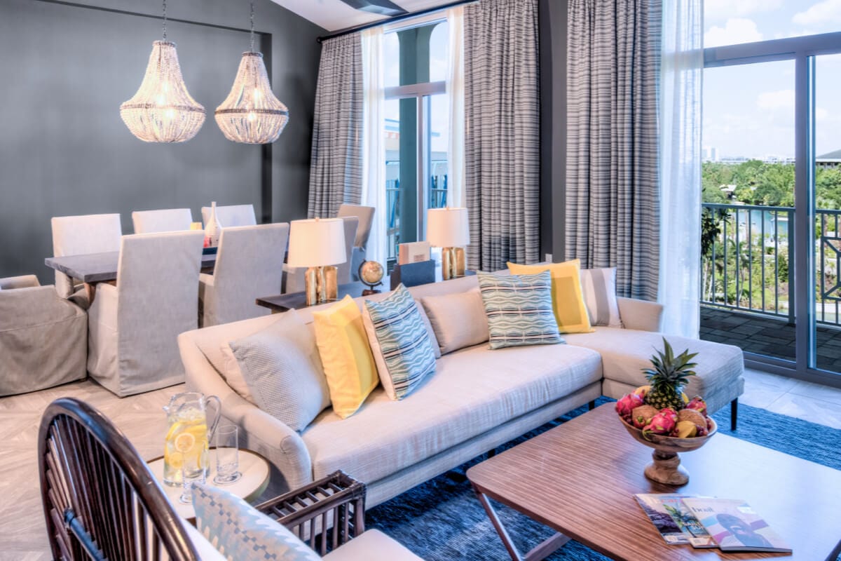 Uma sala de jantar e de estar lindamente mobiliada em uma das suítes do Sapphire Falls Resort Suite apresenta janelas do chão ao teto, mesa de jantar para oito pessoas, sofá seccional e uma tigela de frutas tropicais.