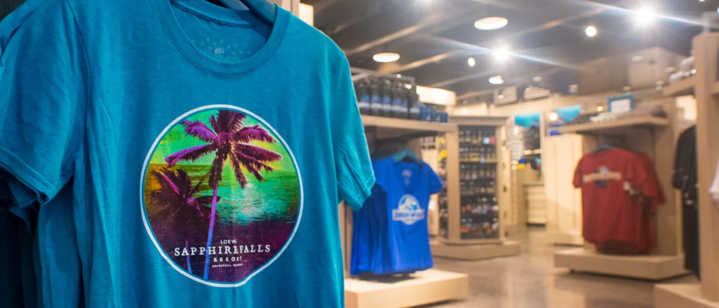 Uma camiseta azul com uma cena de palmeira e oceano com os dizeres "Loews Sapphire Falls Resort" em exposição na loja de presentes do hotel, a Universal Studios Store.