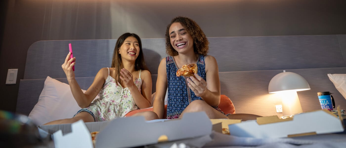 Duas mulheres desfrutando de uma pizza e tirando selfies no quarto do hotel Universal Stella Nova Resort com um papel de parede temático de galáxia ao fundo.