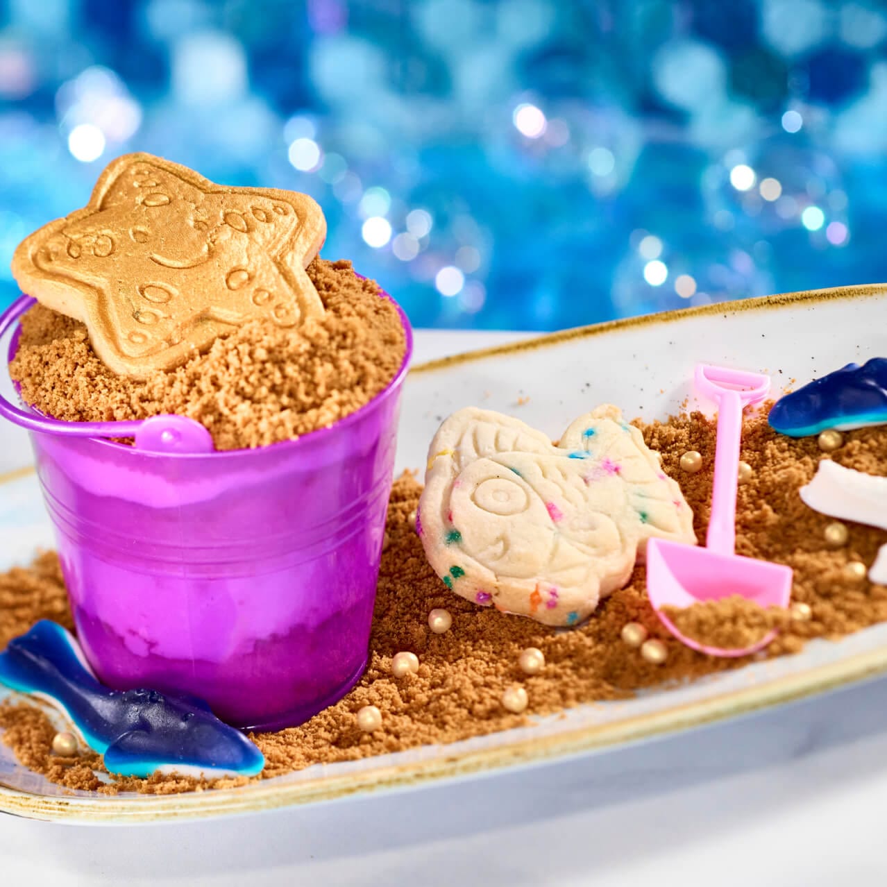 Landlubber's Snickerdoodle Loot com bolo de sickerdoodle em um balde de praia cor-de-rosa, um cookie em forma de peixe, areia comestível e gomas em formato de tubarão, disponíveis no Atlantic.