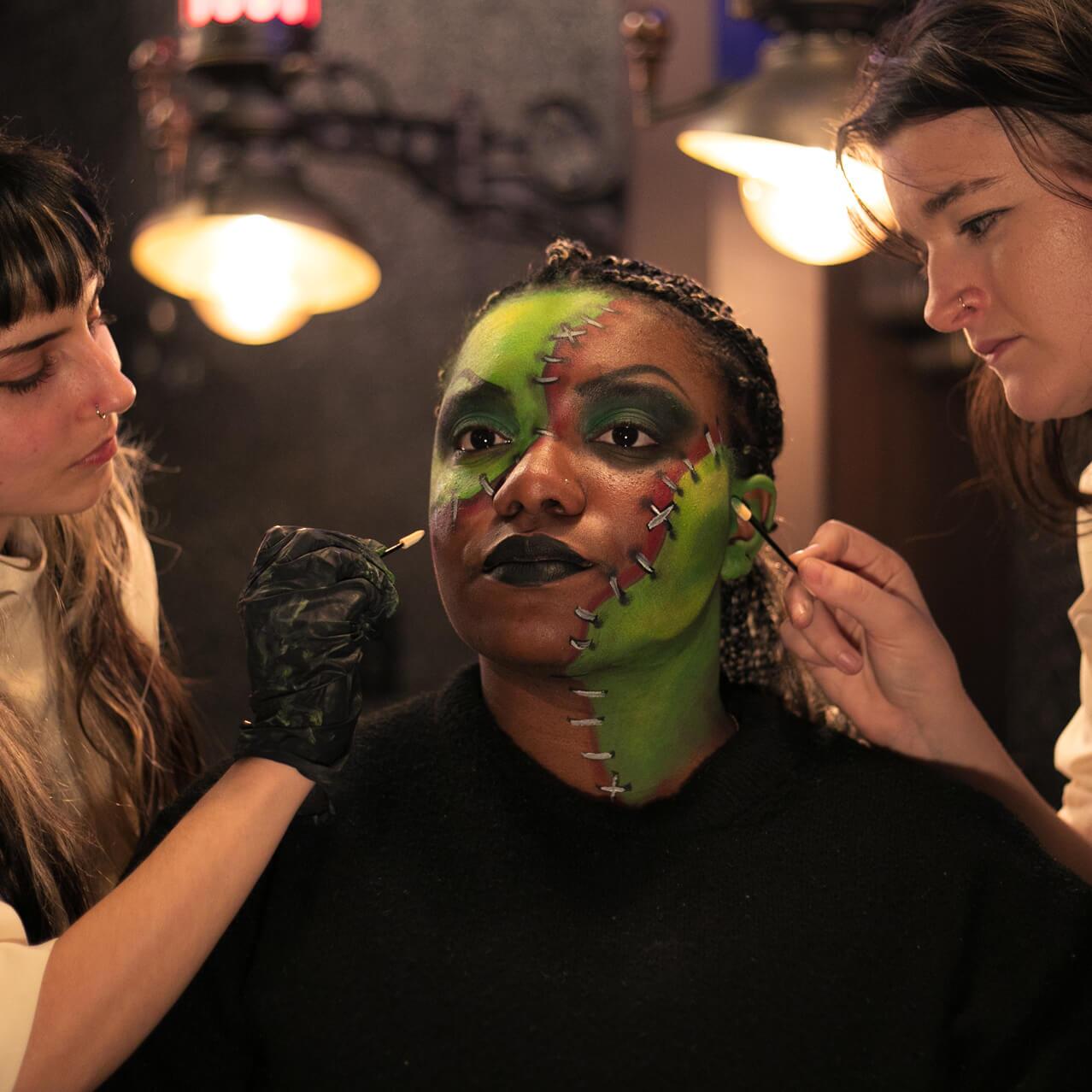 Dois talentosos artistas da Darkmoor Monster Makeup Experience maquiando o rosto de uma cliente, transformando-a no monstro de Frankenstein.