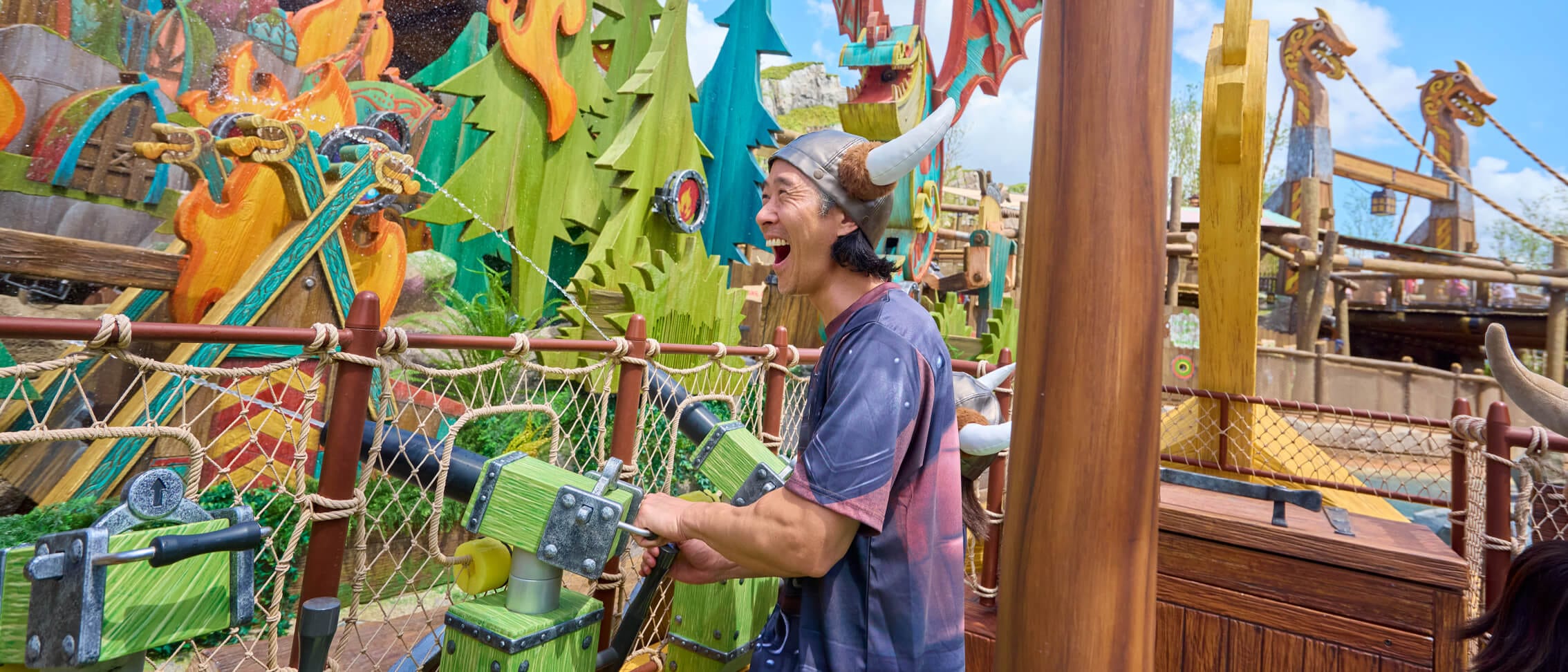 Um visitante usando um capacete viking ri enquanto mira um canhão de água em um alvo do Fyre Drill no How to Train Your Dragon – Isle of Berk.