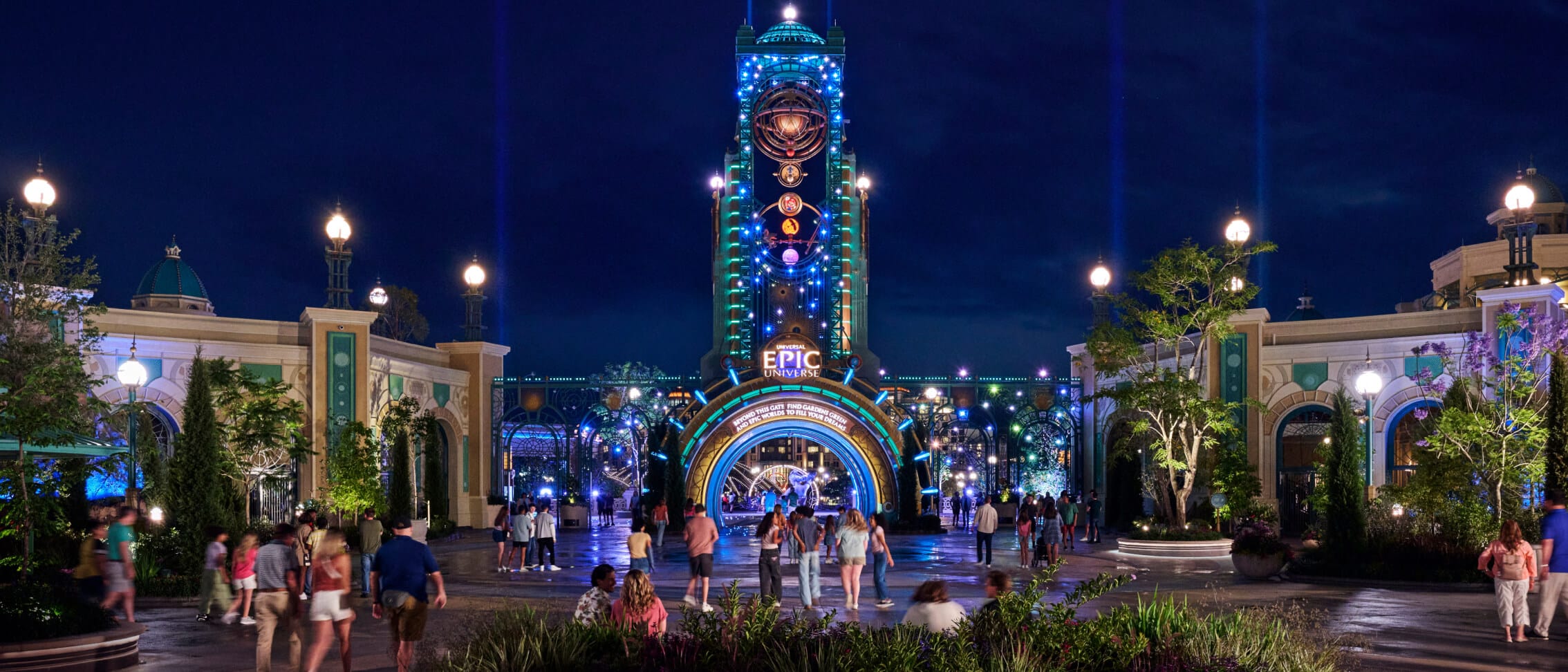 Visitantes caminhando em direção à entrada principal iluminada do Universal Epic Universe, com Cronos surgindo acima sob o céu noturno.