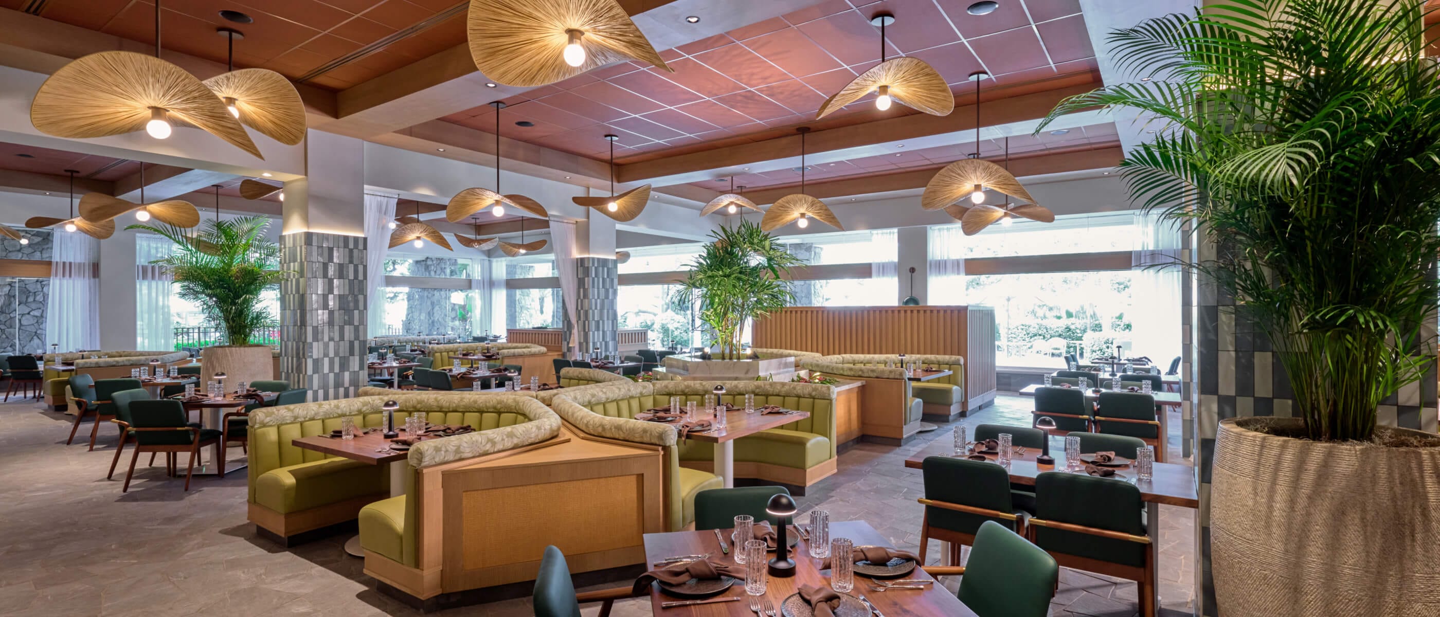 A brightly lit restaurant interior featuring tables and booths with tall plants and fan-style overhead lighting accents.