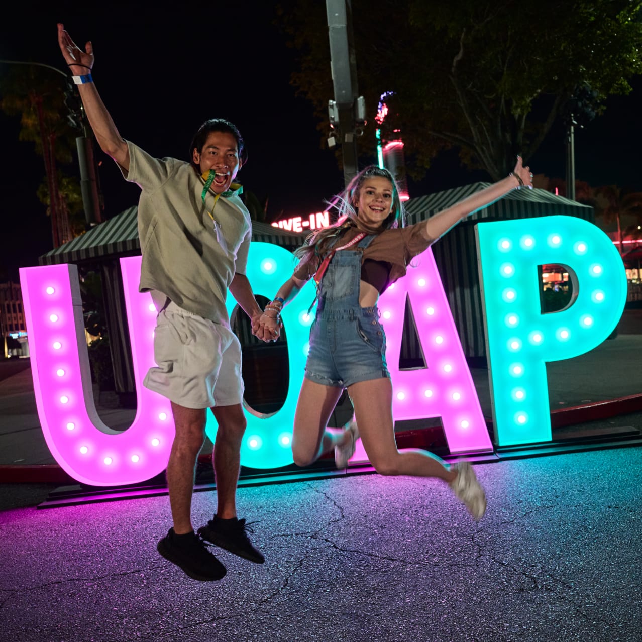 Two excited lanyard-wearing Passholders jumping in the air while holding hands in front of a large “UOAP” sign in pink and green lights.