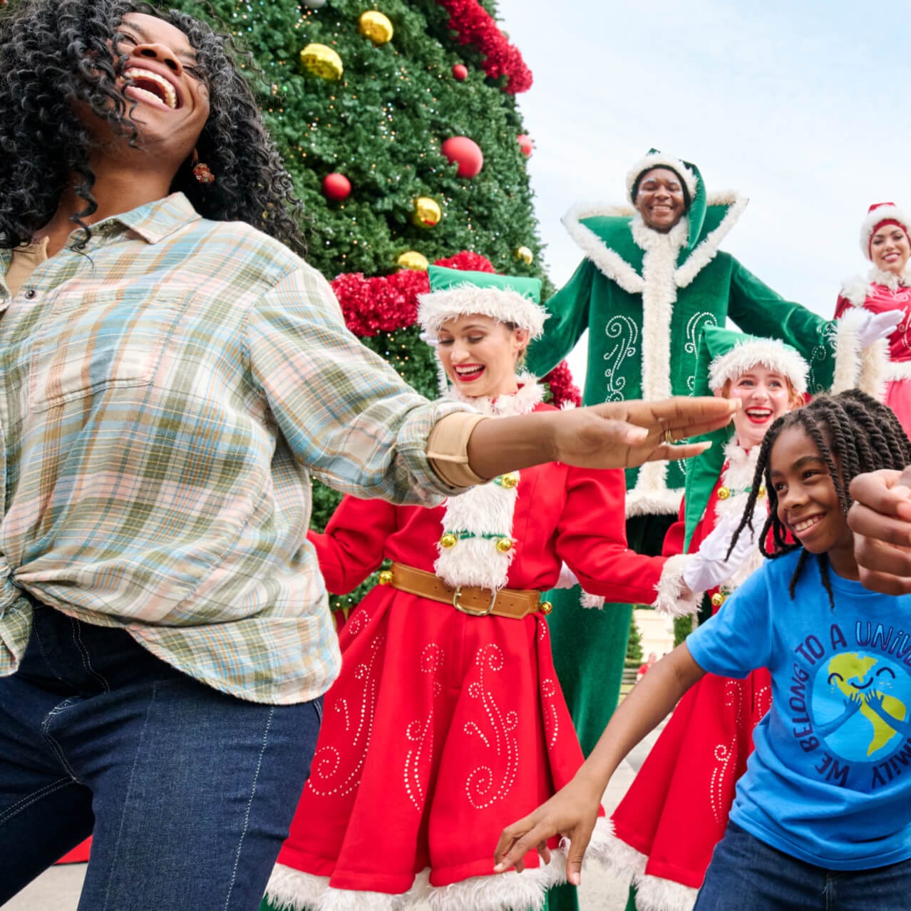Mãe e filhos dançando com elfos vestidos com os tons natalinos vermelho e verde, ao lado de uma grande árvore de Natal no Universal Studios Florida.