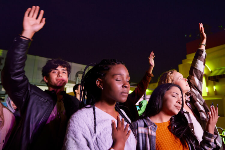 A group of four young adults attending a nighttime concert, some have their eyes closed and several raise their hands in devotion.