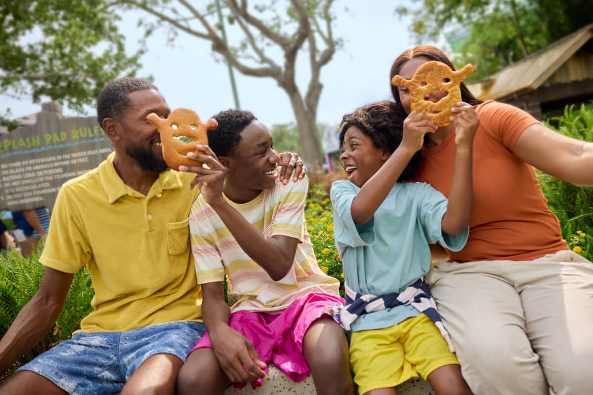 A family on a bench laughing as the kids hold pretzels shaped like Shrek’s face up in front of their parents’ faces.