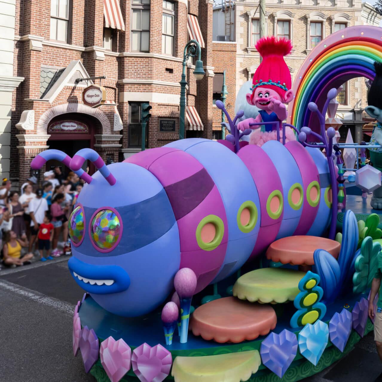 Personagens do filme Trolls em um colorido carro alegórico em formato de ônibus-lagarta enquanto visitantes assistem da calçada.