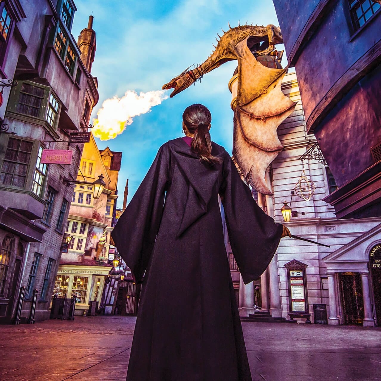 As lojas do Diagon Alley™ se erguem de ambos os lados, enquanto uma garota vestindo uma capa de Hogwarts™ com uma varinha olha para um dragão que cospe fogo no topo do banco Gringotts™ no Universal Studios Florida.