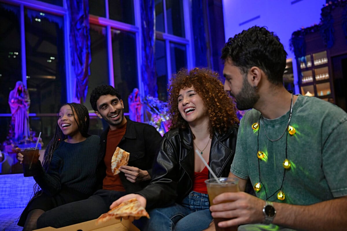 A group of excited friends enjoying pizza and drinks with glowing purple décor behind them.