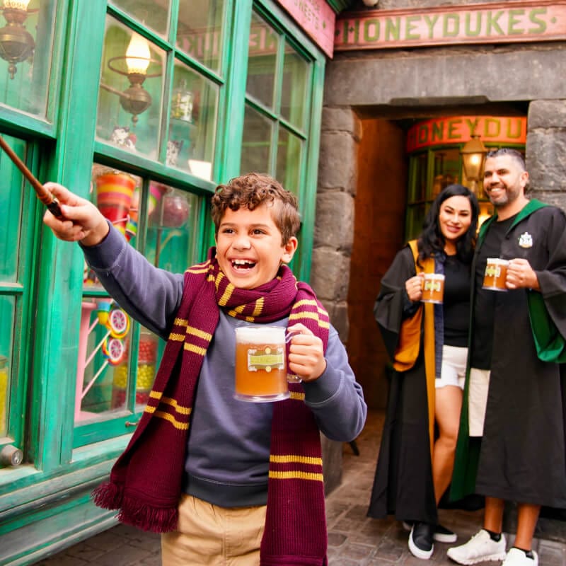 Uma família de três pessoas se diverte tomando Butterbeer™, e os pais observam o filho erguer sua varinha.