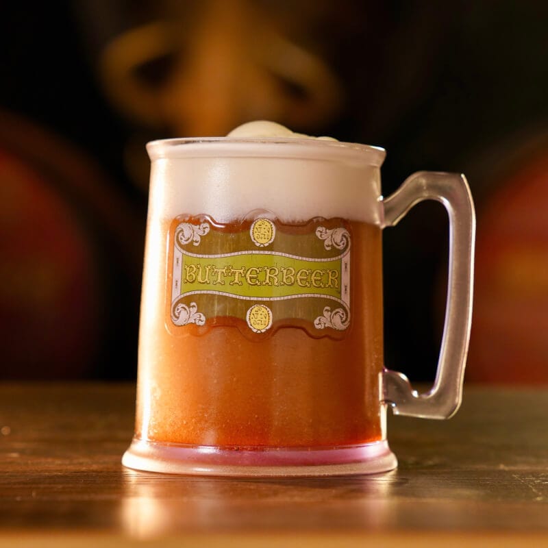 Uma caneca transparente com alça e o logo “BUTTERBEER™", cheia até a borda, apoiada sobre uma superfície de madeira em um ambiente de bar com iluminação baixa.