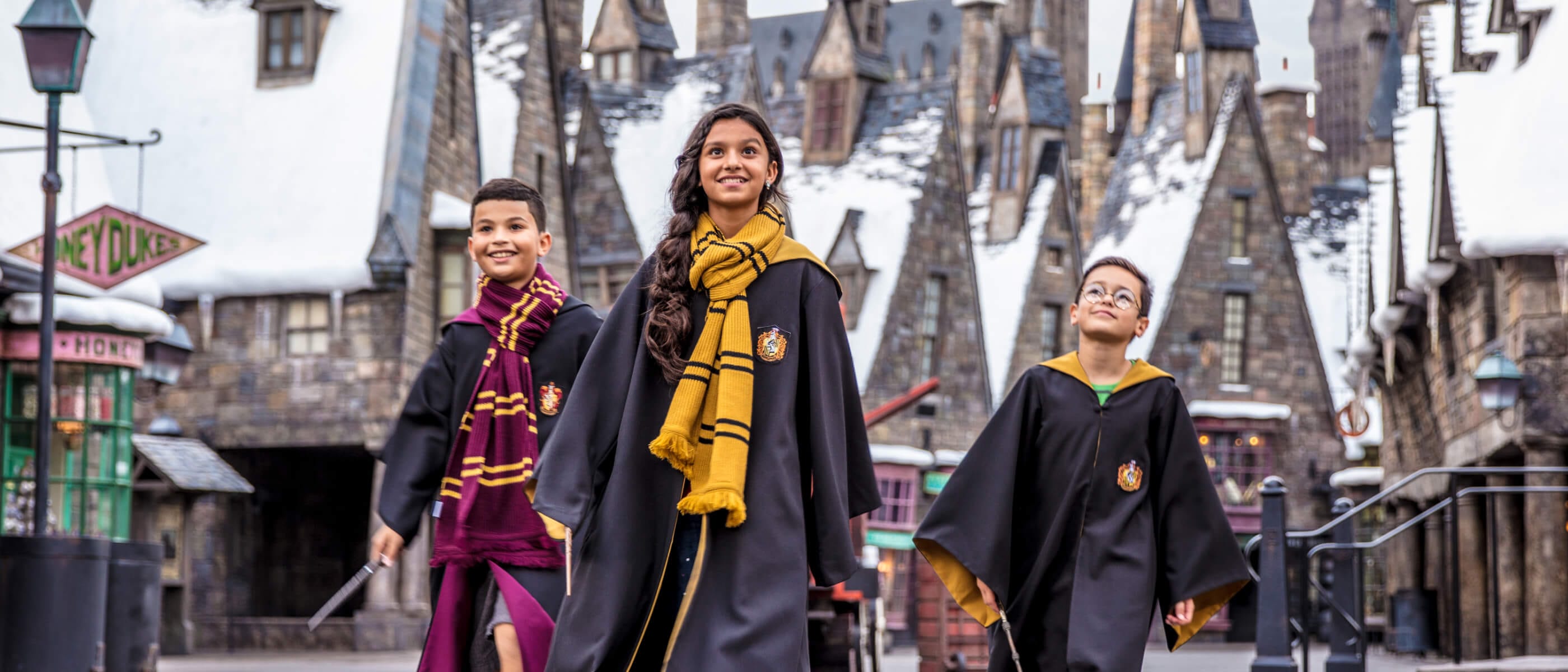 Wearing wizard robes and carrying wands, three children walk together through Hogsmeade village at Universal’s Islands of Adventure.
