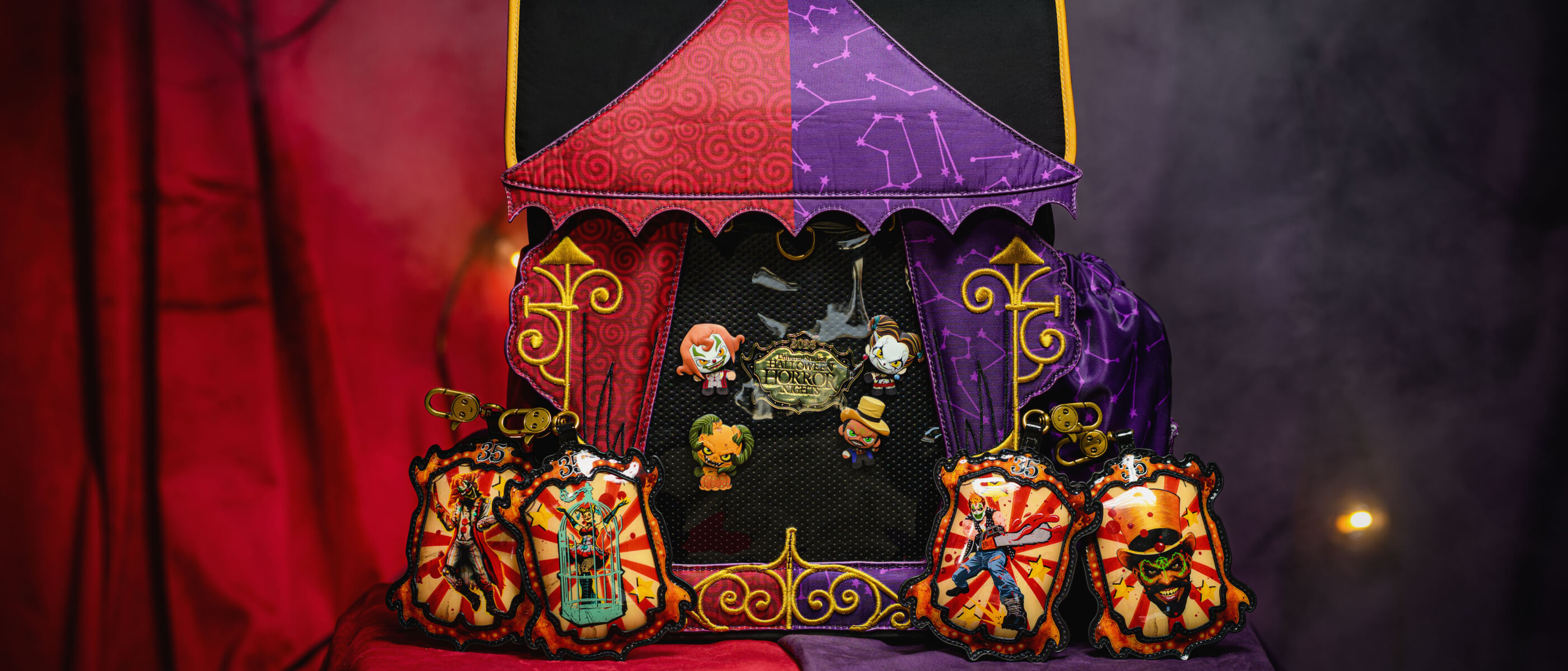Exposição de produtos comemorativos do aniversário de 35 anos do Halloween Horror Nights, com uma mochila em formato de tenda de circo vermelha e roxa e chaveiros.