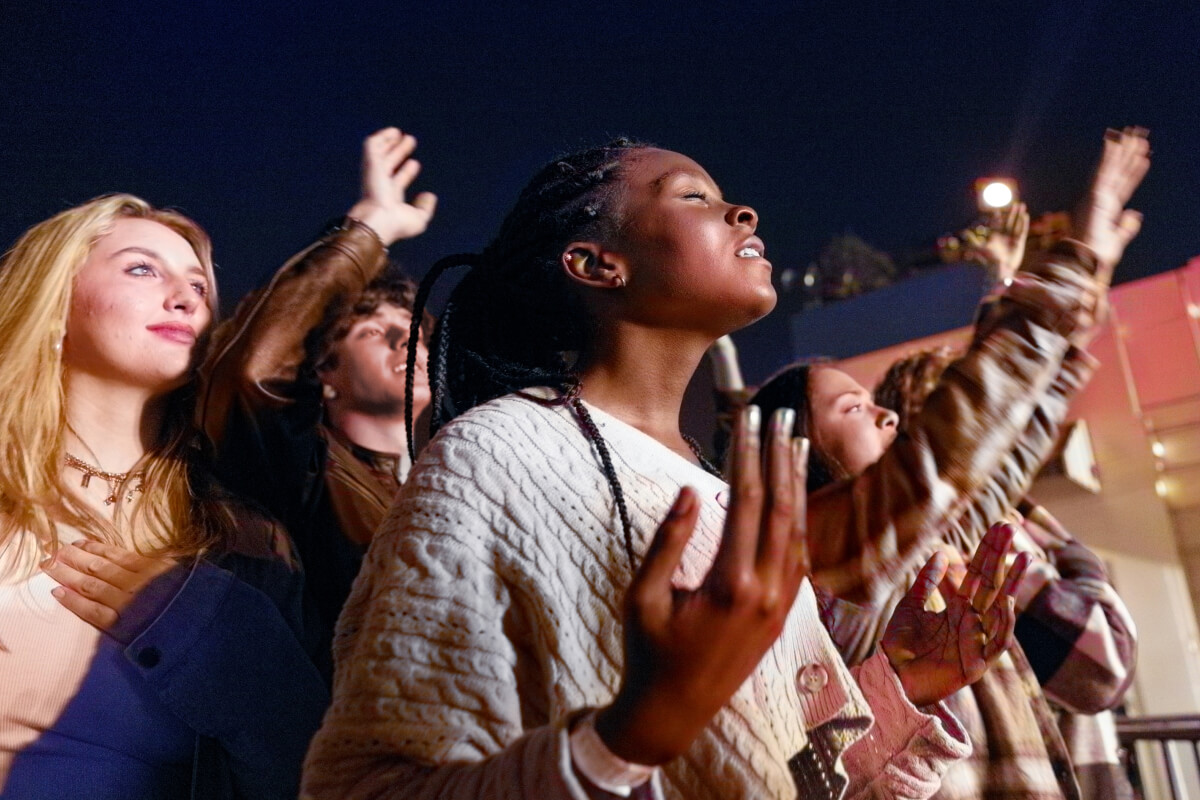A group of five young adults attending a nighttime concert, some have their eyes closed and several raise their hands in devotion.&nbsp;