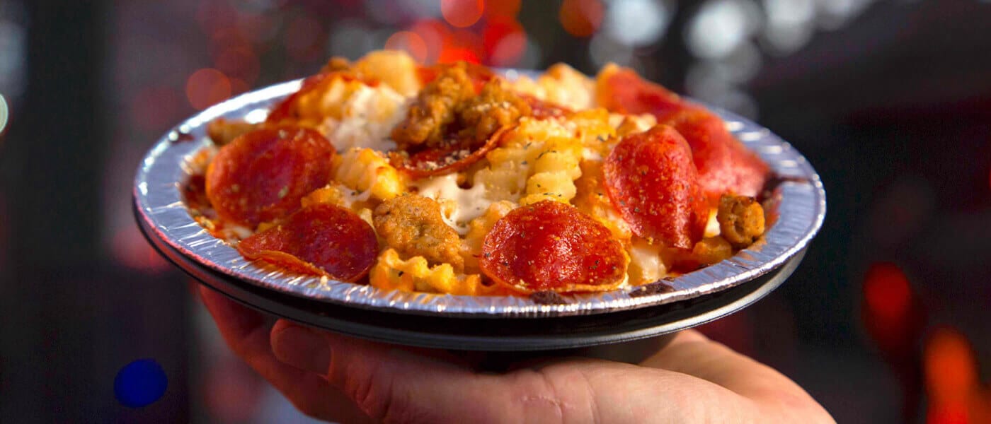 A hand holding a bowl of crinkle cut fries topped with pepperoni, cheese and pieces of breaded chicken.