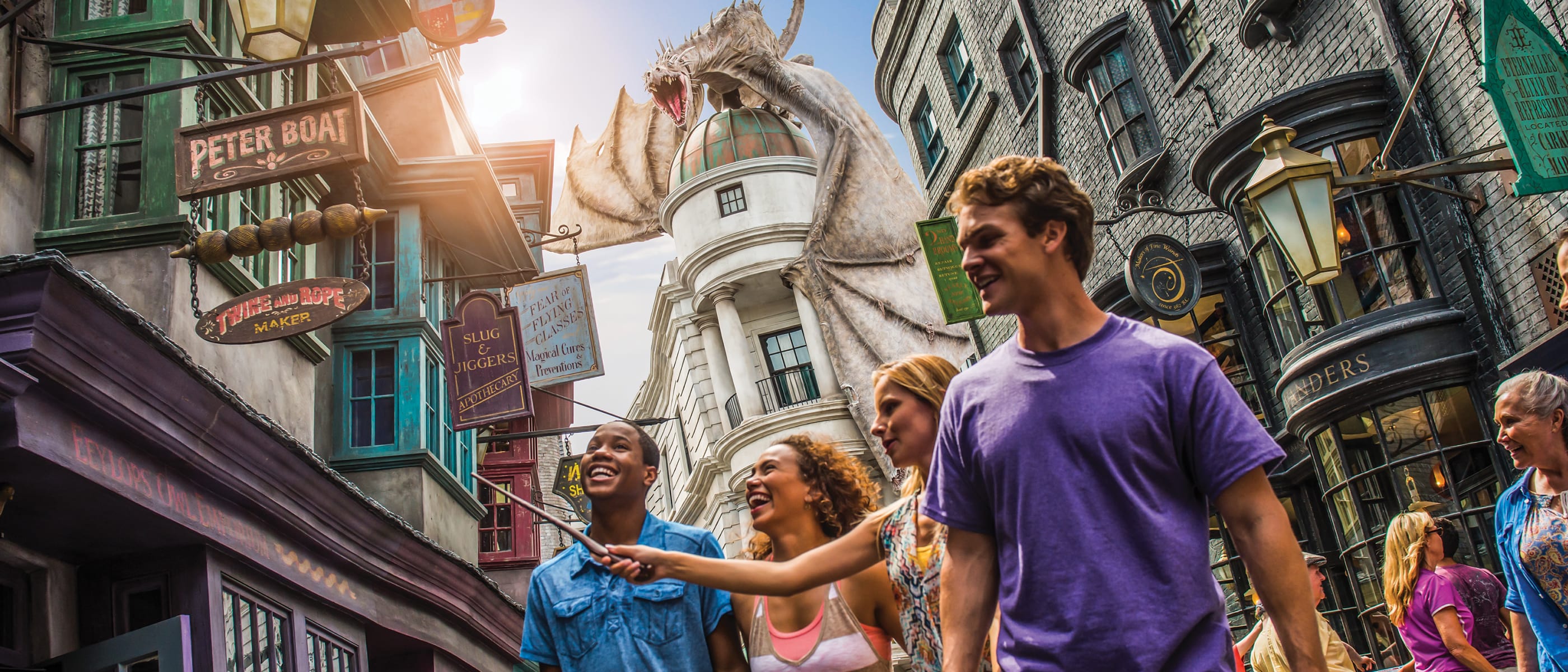  Un grupo de amigos sonriendo mientras uno apunta con una varita a un edificio en Diagon Alley con el dragón de Gringotts arriba