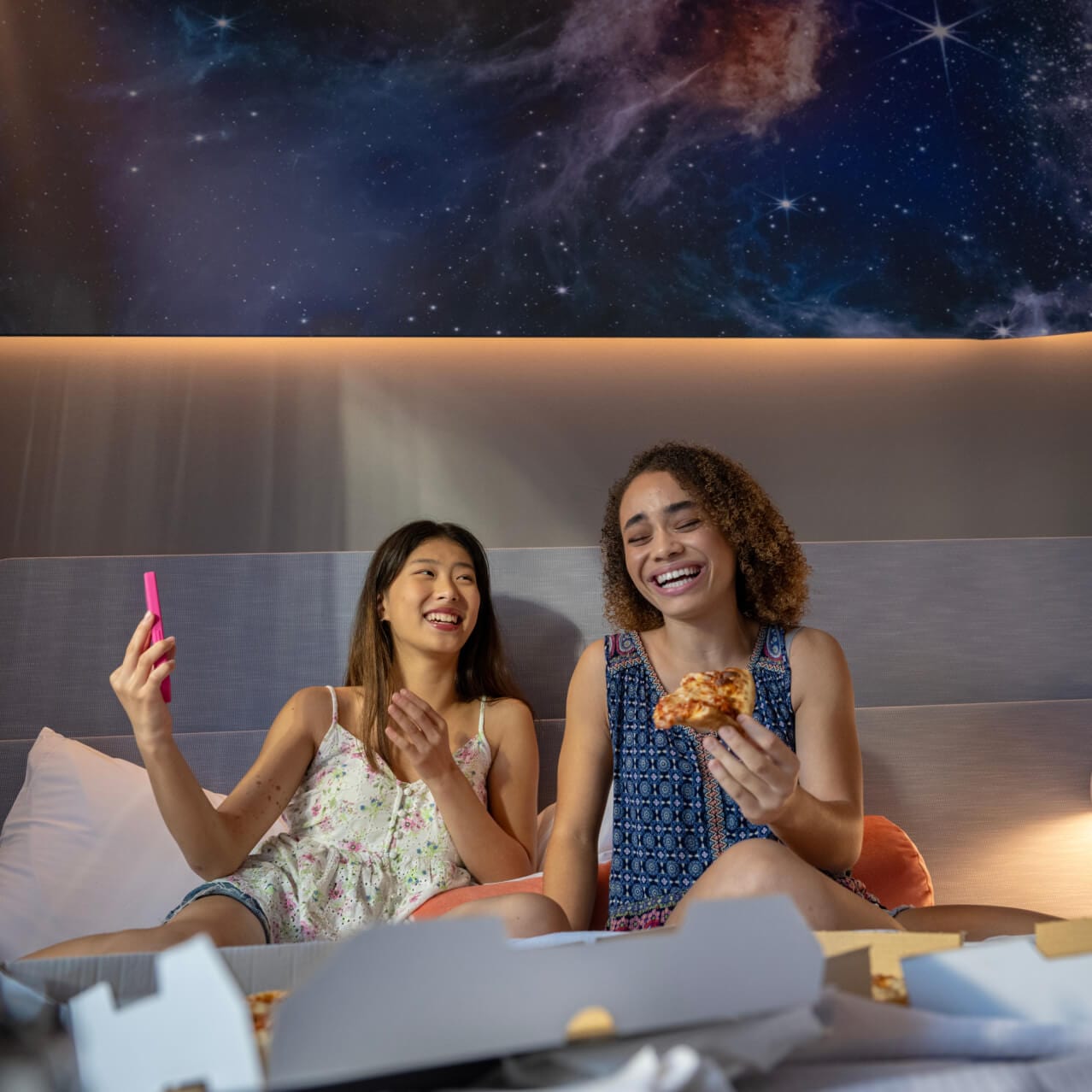 Two women enjoying pizza and selfies in their Universal Stella Nova Resort hotel room with a galaxy themed wallpaper in the background.