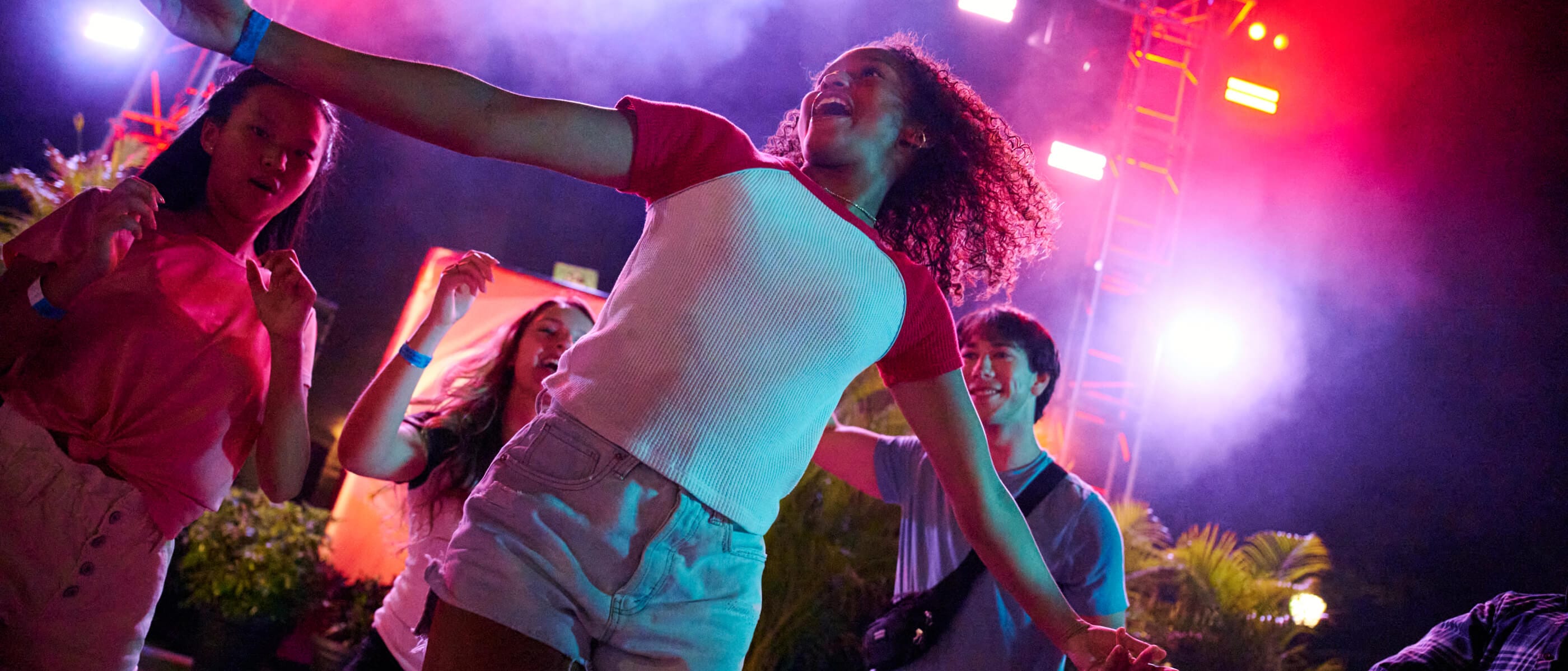A group of young, excited Guests dancing in front of a stage with colored lights and fog at night.