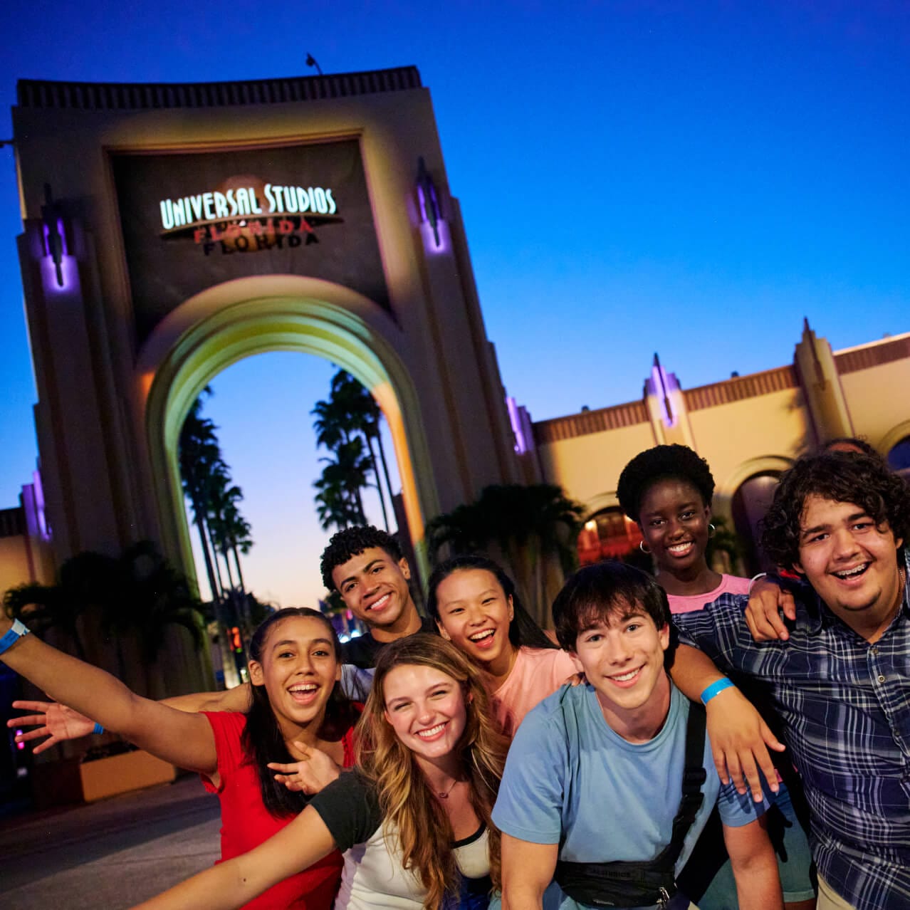 A group of young smiling Guests posing for a photo together in front of the Universal Studios Florida entrance arch at night.