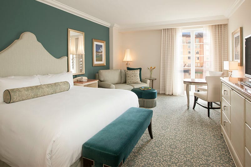 The interior of a King Guest Bedroom in Loews Portofino Bay Hotel at Universal Orlando, complete with white linens, royal turquoise accents, and lush furniture and decor. 