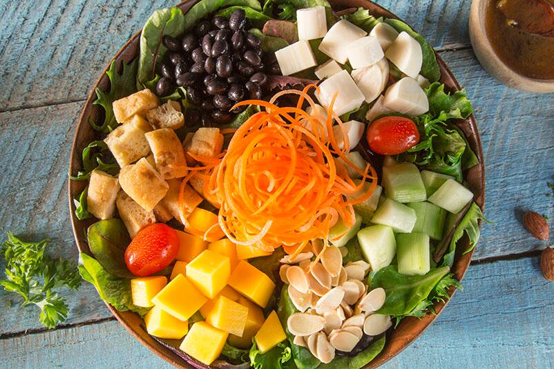 A photo of Tropical Baby Greens topped with shaved carrots, hearts of palm, diced mango, grape tomatoes, cucumbers, tofu, black beans and toasted almonds with soy garlic vinaigrette at Universal's Volcano Bay.
