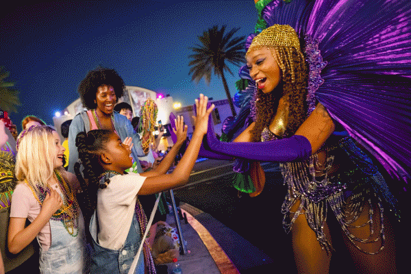 A GIF including a child high fiving a performer, a family dancing along, guests wearing beads and friends watching a performance at Universal Mardi Gras.