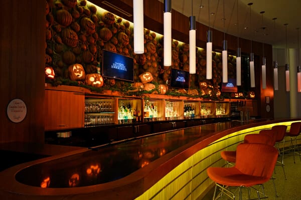 A stylish, low-lit bar with high-top seating and a series of carved pumpkin decorations all along its back wall.