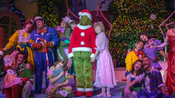 The Grinch dressed up as Santa and making a mischievous facial expression with Seuss Landing decorated with Christmas lights in the background.
