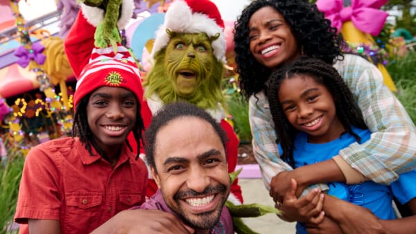 Selfie of a family smiling next to the Grinch at Seuss Landing™ during Grinchmas™ at Universal Islands of Adventure.Adventure.Seuss Landing decorated with Christmas lights in the background.